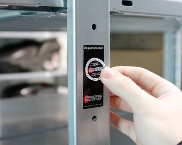Plakette zur Regalinspektion wird an Metallregal angebracht – dokumentierte Prüfung für sichere und vorschriftskonforme Lagertechnik.